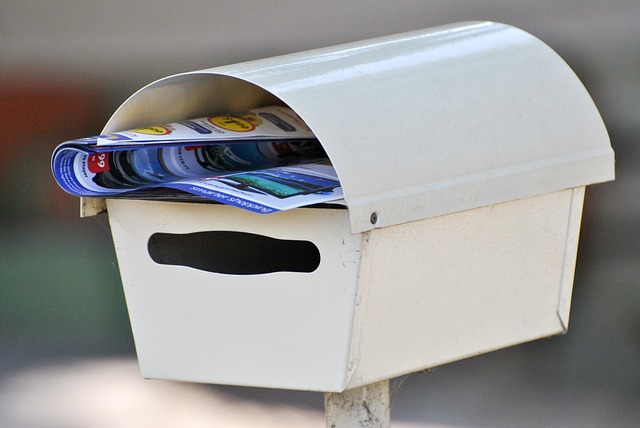 A black metal letterbox with a mail slot and lockable compartment mounted on a wooden fence.